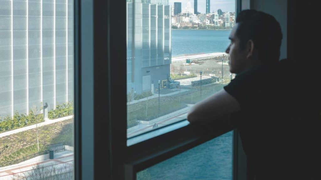 A man looks out a window at a body of water and city buildings.