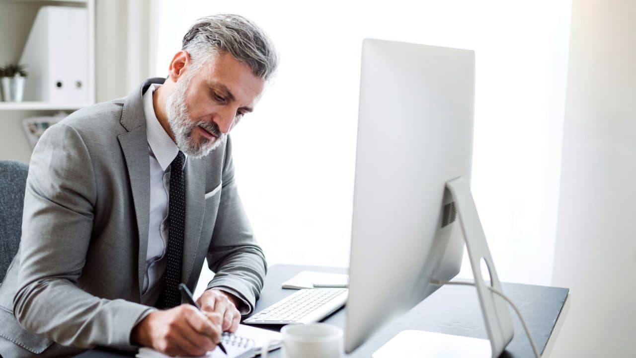 A bearded man in a suit writes in a notebook at a desk with a computer.