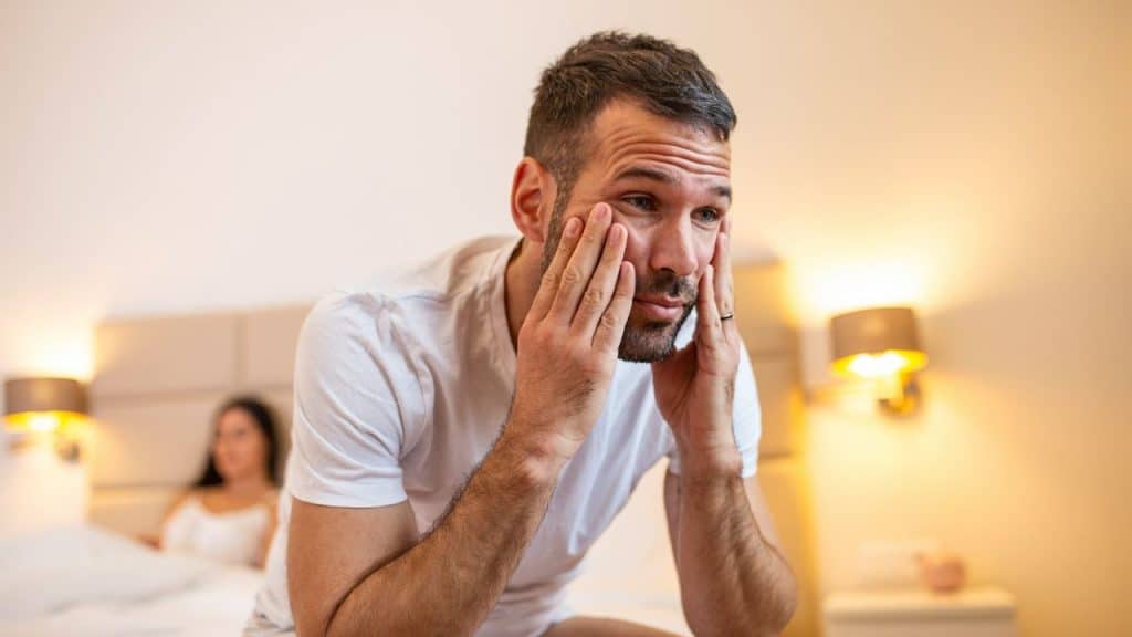 A distressed man with hands on his face sits on a bed, while a woman lies in the background.