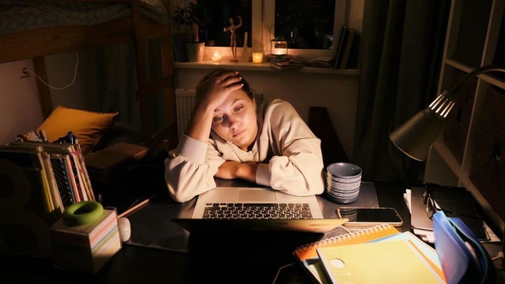 A distressed woman sits at a cluttered desk with a laptop, head in hand.