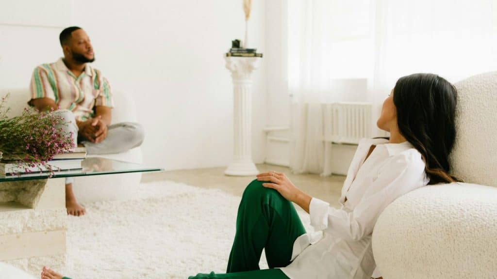A woman sits on the floor looking up at a man sitting on a sofa in a bright room.