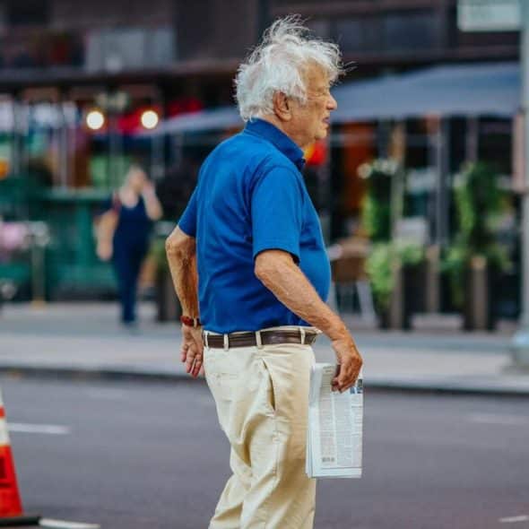 A senior man with white hair, wearing a blue polo shirt, walks across a street.
