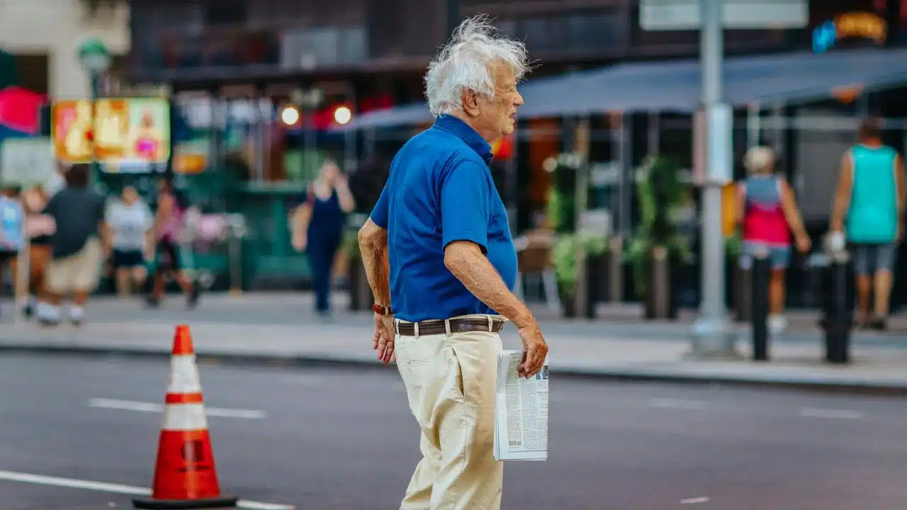 A senior man with white hair, wearing a blue polo shirt, walks across a street.