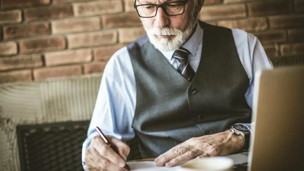 An older man in a vest writing at a desk beside his laptop.