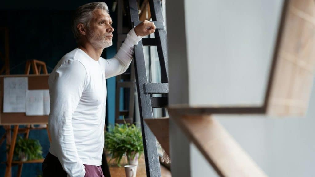 A serious man with gray hair and a beard leans against a dark metal structure.