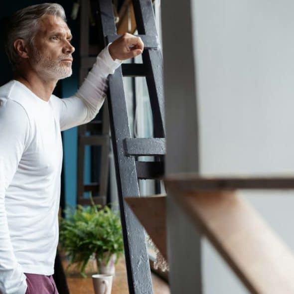 A serious man with gray hair and a beard leans against a dark metal structure.