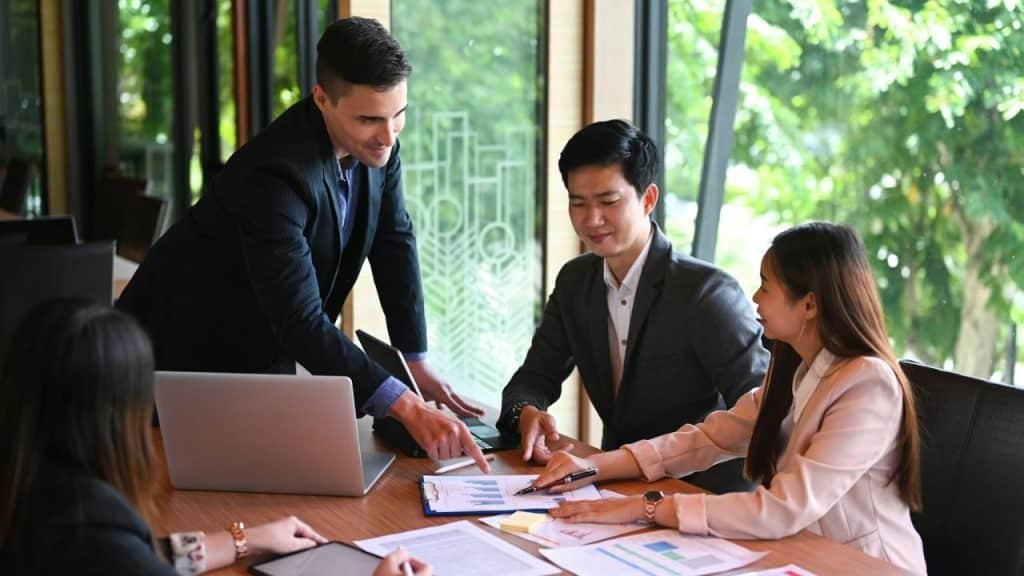 A smiling man in a suit points to a document on a table, surrounded by three other people.