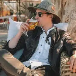 A man in a safari hat and leather jacket sipping beer outdoors.