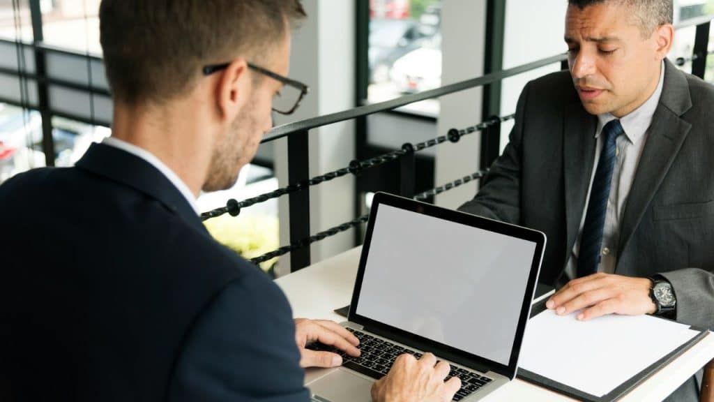 A man in glasses types on a laptop across from another man in a suit.