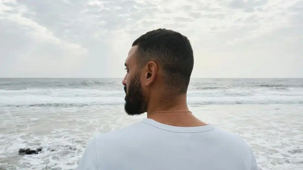 An adult man with short hair and a beard looking at the ocean under a cloudy sky.