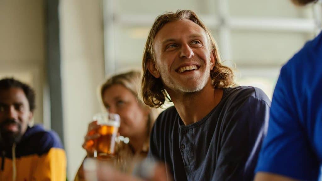 A smiling man holding a beer while chatting with friends.