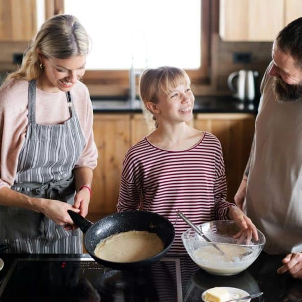 A family cooking pancakes together in a bright wooden kitchen.