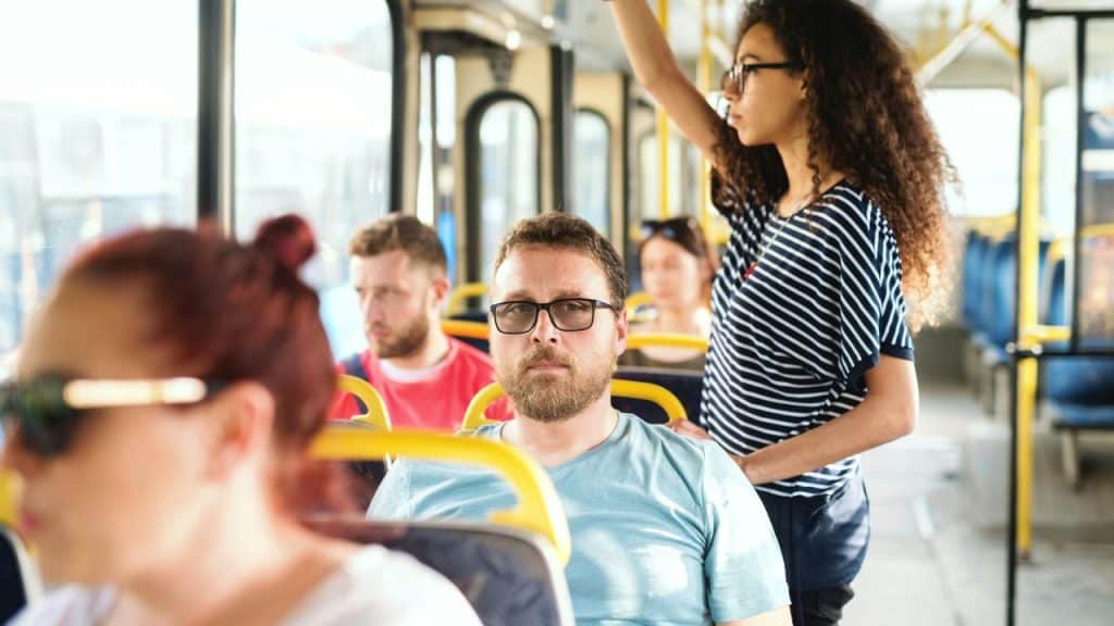 A man with glasses seated on a bus with a standing passenger behind him.