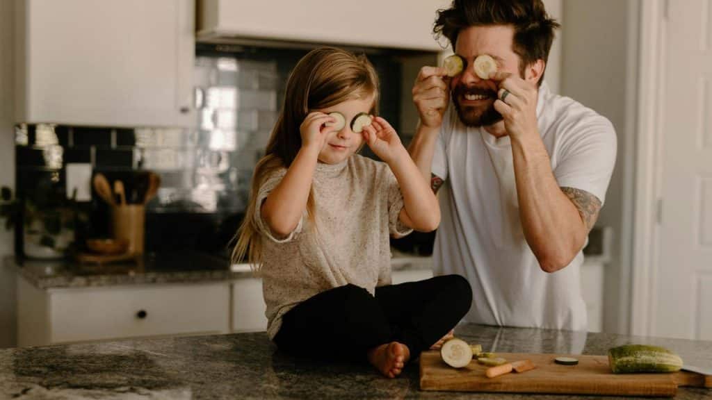 A man and a girl playfully holding cucumber slices over their eyes in a kitchen.