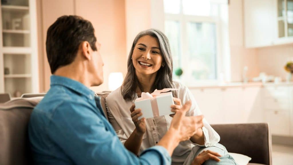 A woman offering a wrapped gift to a man on a couch.