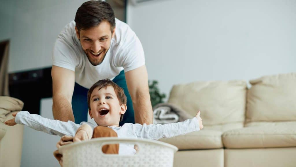 A man pushing a smiling child seated in a laundry basket.