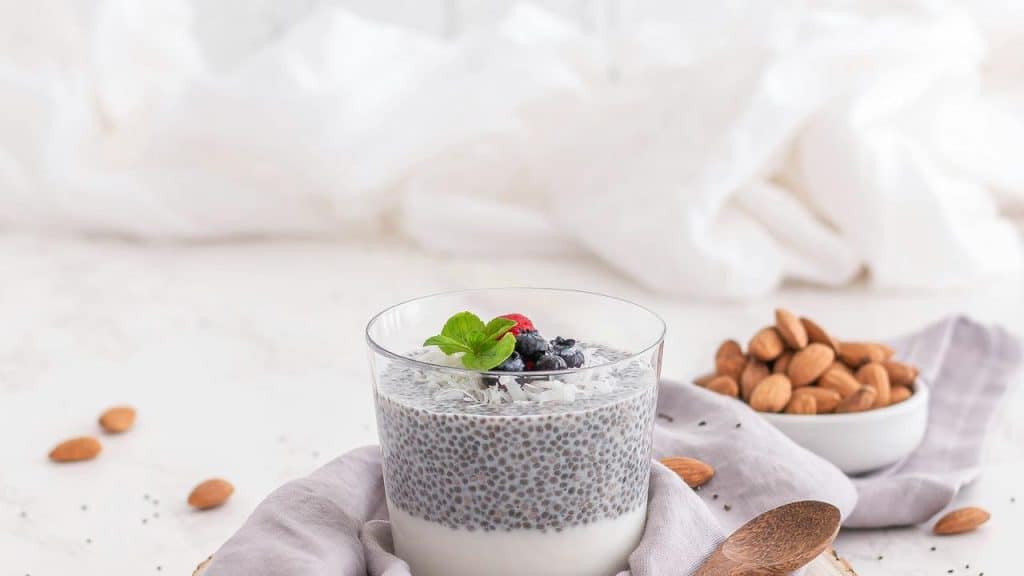 A glass of chia pudding topped with berries, coconut flakes, and mint beside a bowl of almonds.