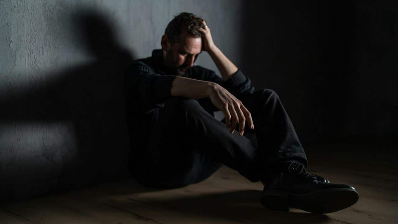 A man sits on a floor in a dark room, head in his hand, looking down.