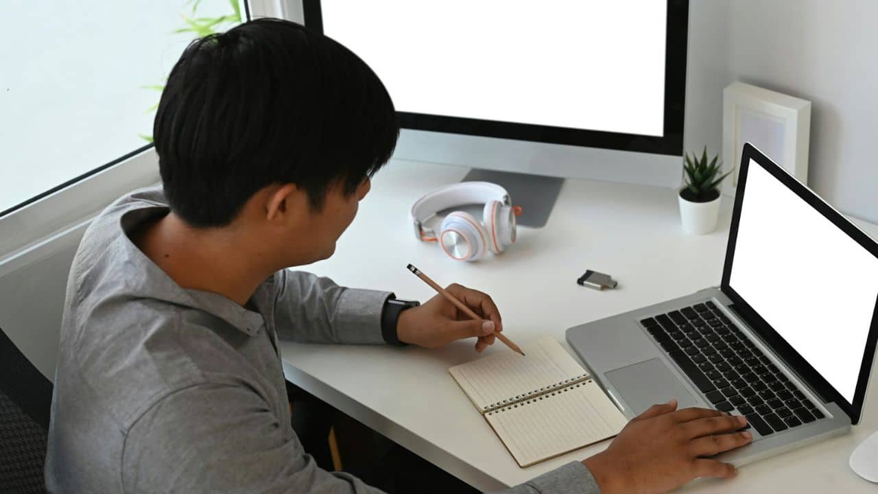 A person works at a desk with a laptop, monitor, and notebook.