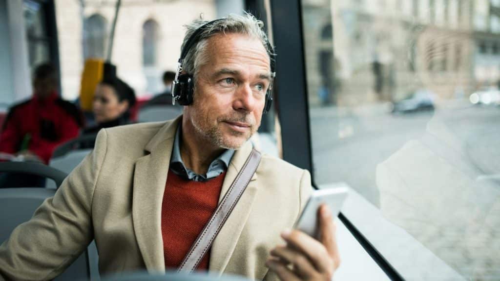 A gray-haired man with headphones looks out a bus window, holding a phone.