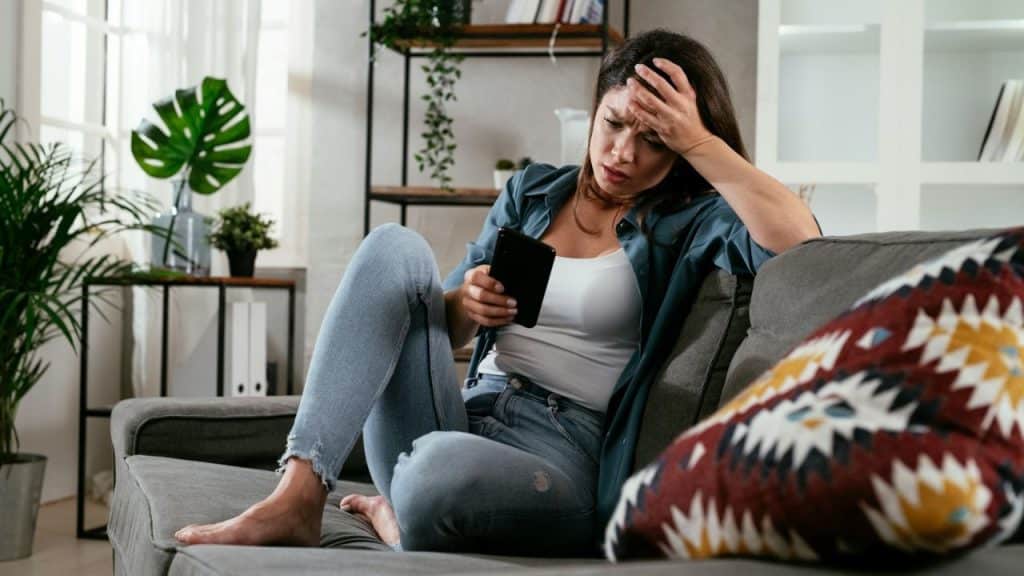 A frustrated woman sits on a couch, looking at her phone with her hand on her forehead.
