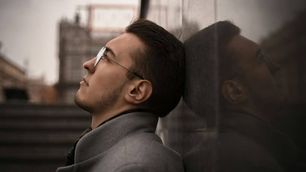 A man in a grey coat and glasses leans against a dark, reflective surface, looking upwards.
