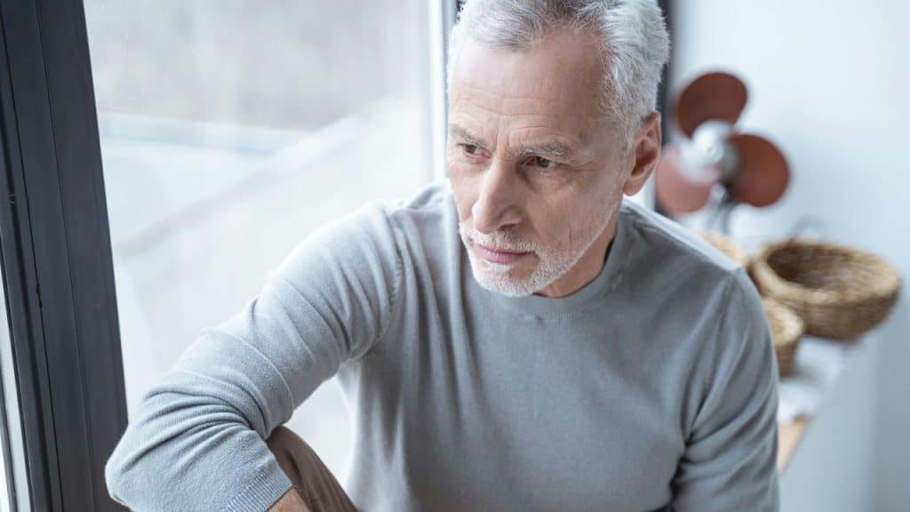 A contemplative older man with gray hair and a beard looks out a window.