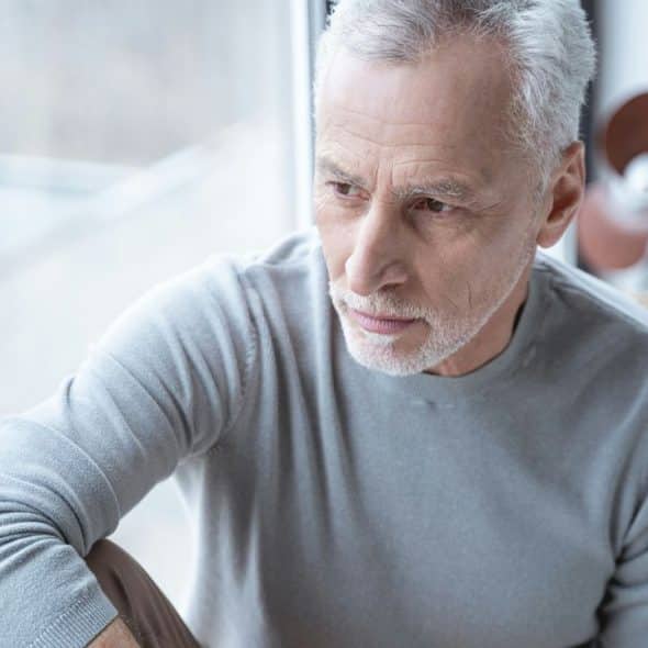 A contemplative older man with gray hair and a beard looks out a window.