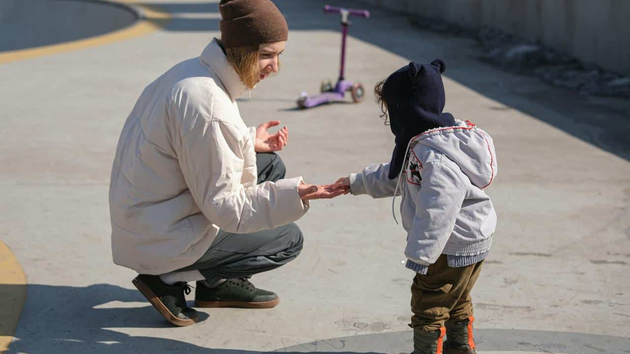 An adult crouching and offering their hand to a toddler outdoors.