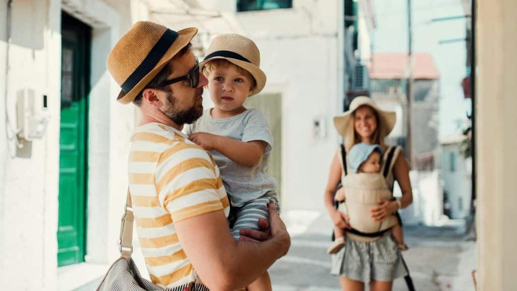 A father holding a toddler while mother carries a baby in a carrier on a sunny street.