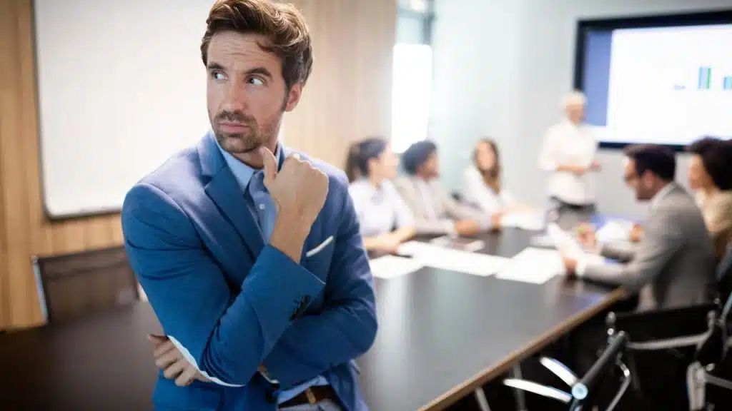 A pensive man in a suit standing before a conference table with colleagues meeting behind him.