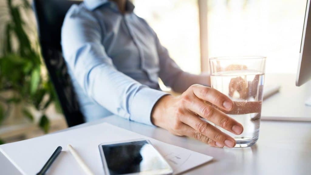 A person's hand reaches for a glass of water on a desk next to a smartphone and papers.