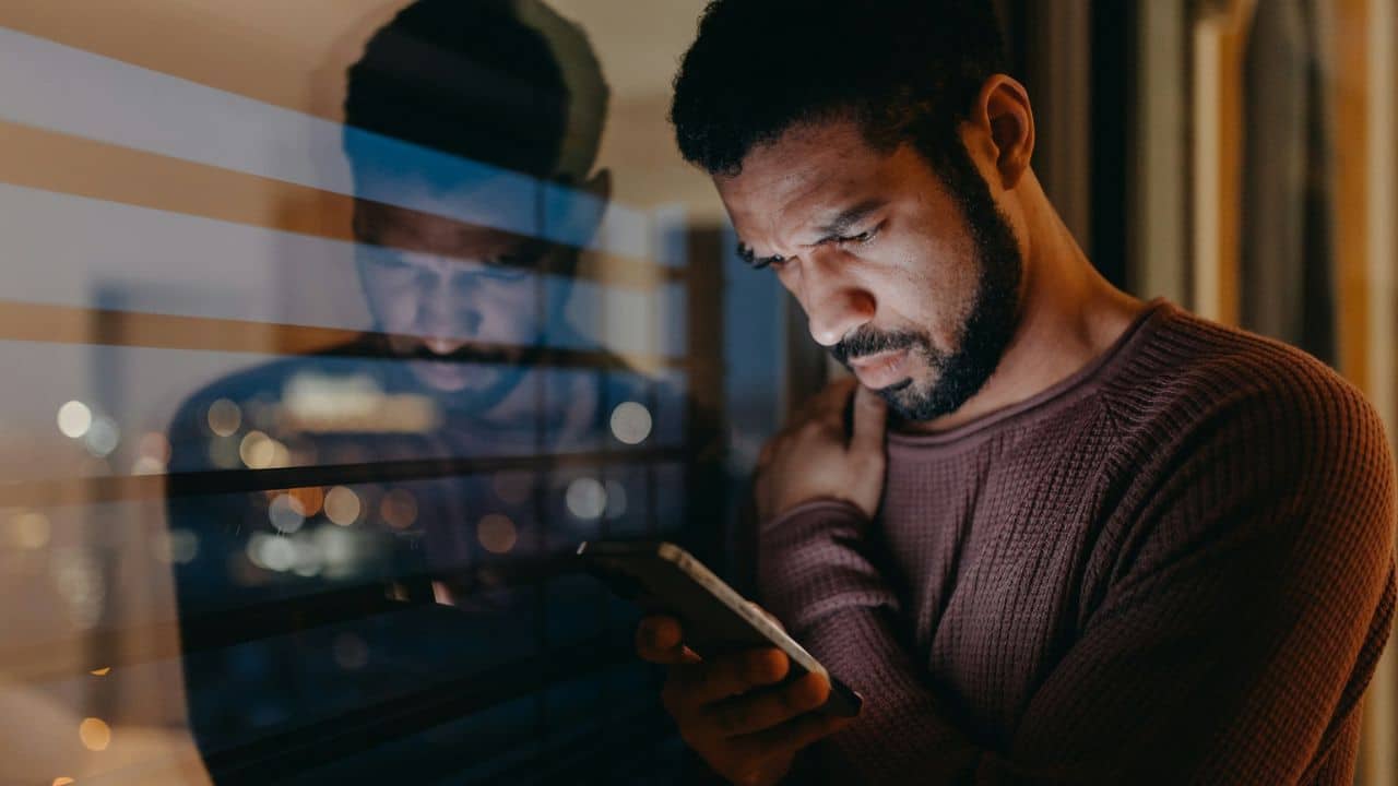 A man with a beard looks intensely at his phone, with his reflection visible in a window.