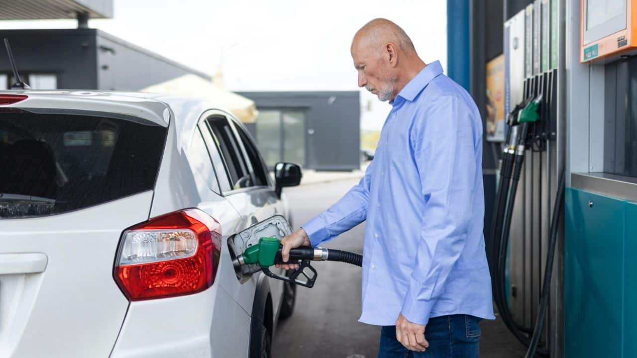 A man in a blue shirt is filling a white car with gas at a pump.