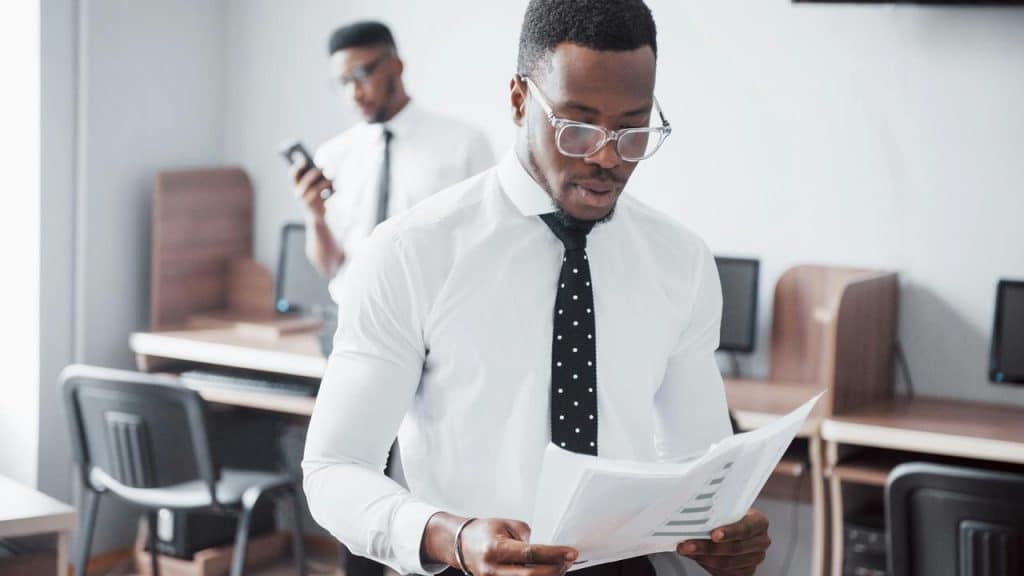 A man in glasses reading papers at his office desk with a colleague behind.