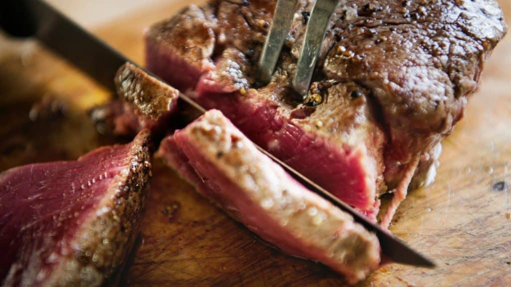 A knife slicing a medium-rare steak on a wooden cutting board.