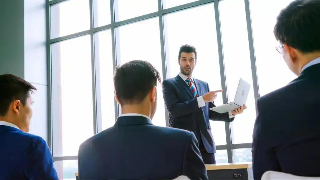 A man in a suit presenting to colleagues with a laptop.