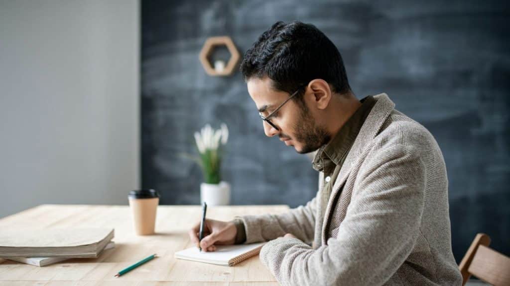 A man with glasses and a beard writes in a notebook at a wooden table.