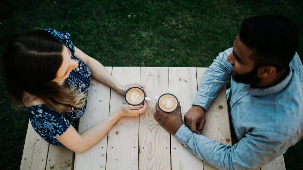 A top-down view of two people holding coffee mugs at a wooden table outdoors.