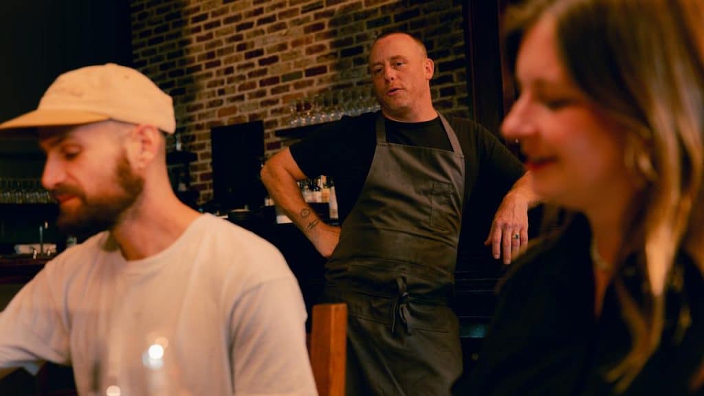 A chef standing behind diners at a restaurant table.