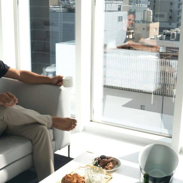 A man sitting on a couch holding a mug and gazing out a sunlit window.