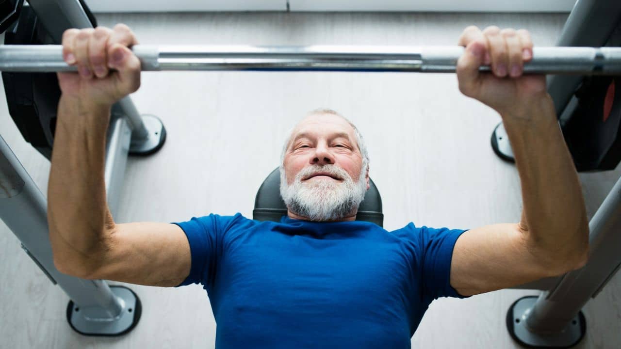 A smiling, bearded man in a blue shirt performs a bench press.