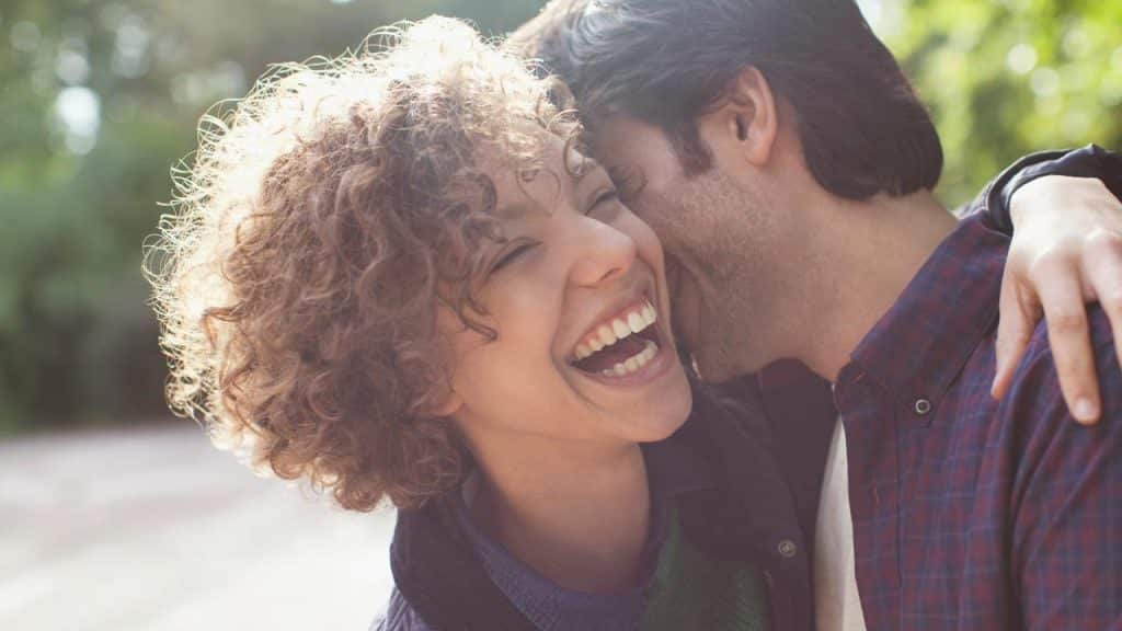 A curly-haired woman laughing as a man whispers in her ear outdoors.