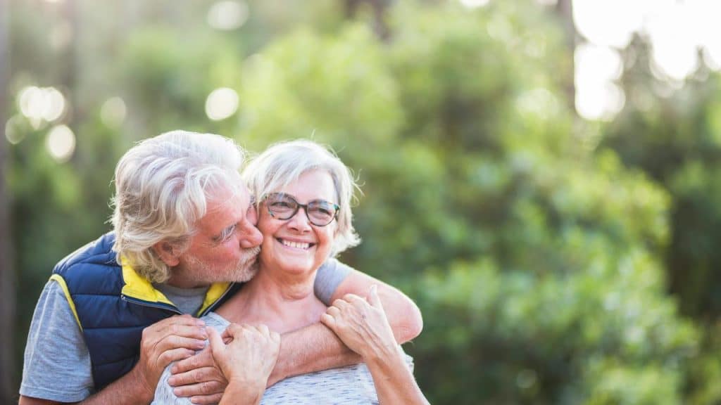 An older man kissing his smiling partner on the cheek outdoors.