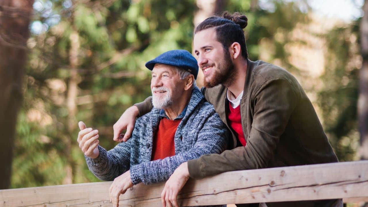 A cheerful older man and younger man stand together outdoors, looking off into the distance.