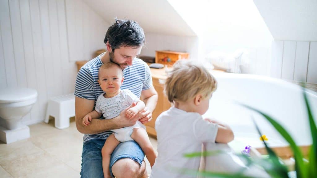 A father holding a crying baby as a toddler plays at the bathtub.