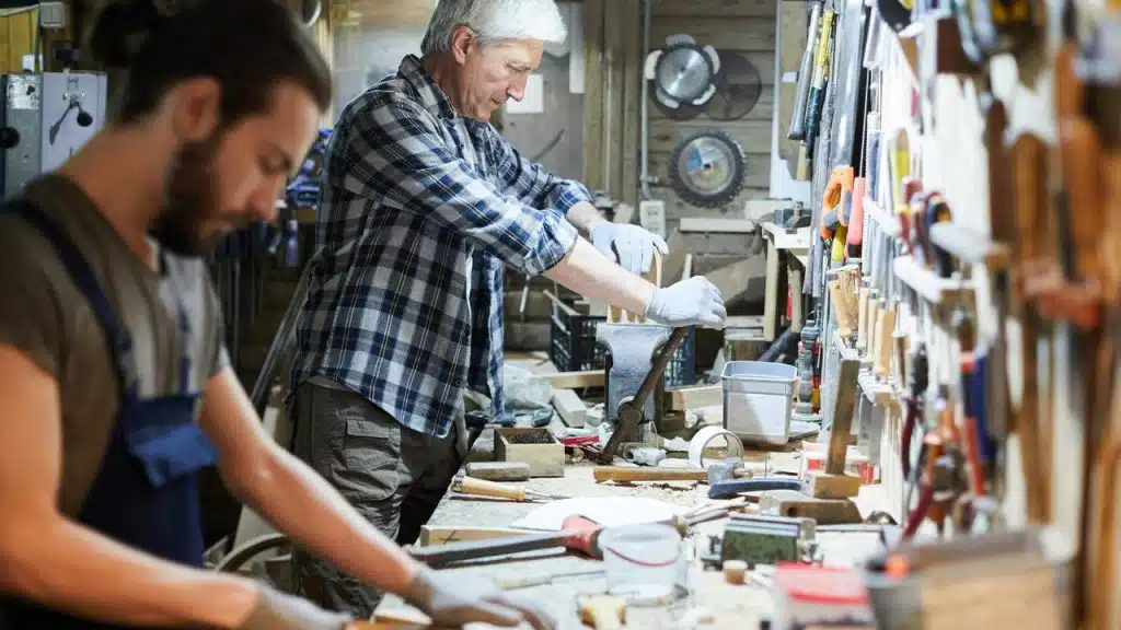 A senior woodworker and his apprentice working at a cluttered bench.