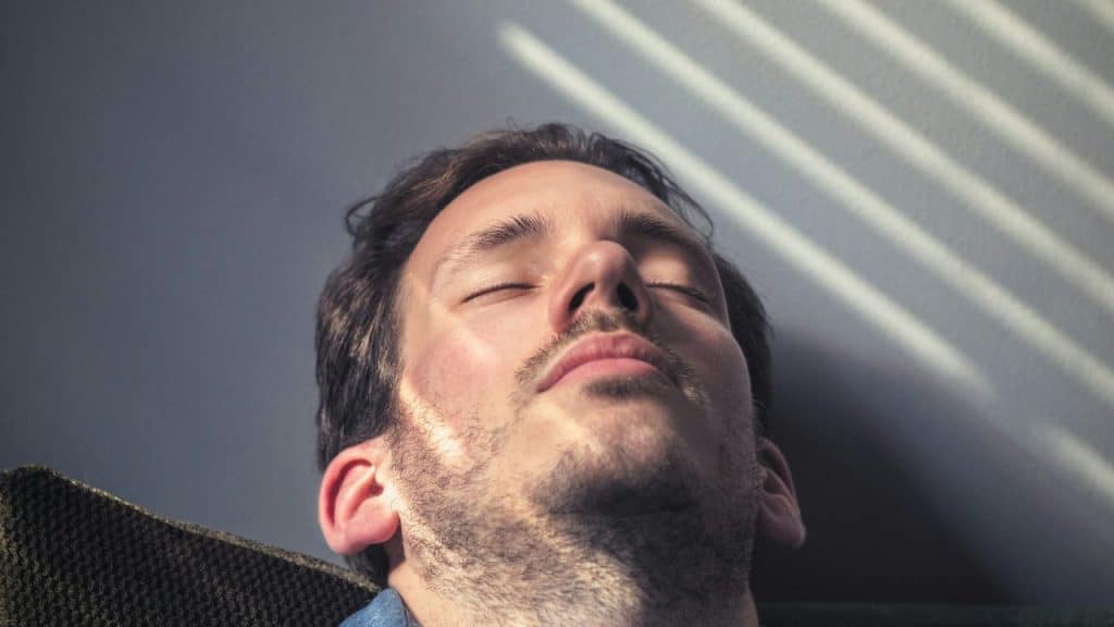 A man with his eyes closed rests his head against a wall with striped shadows.