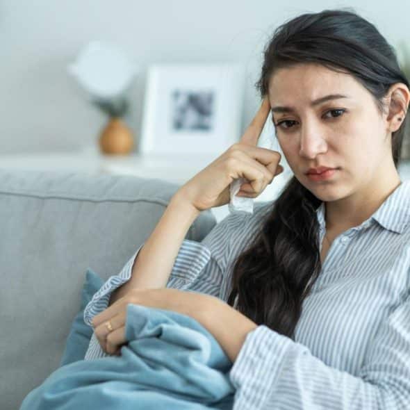A young woman sits on a couch, holding her head, with a pensive expression.