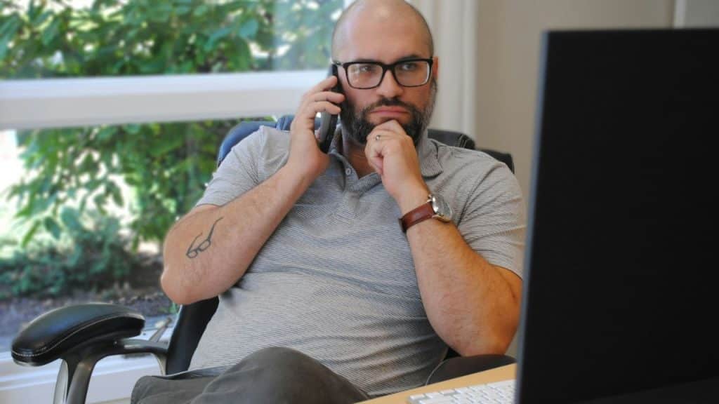 A man with glasses and a beard talks on a phone, looking at a computer screen.